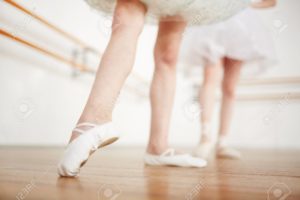 Faceless girls leg in ballet shoes making position on parquet with ballerina on background